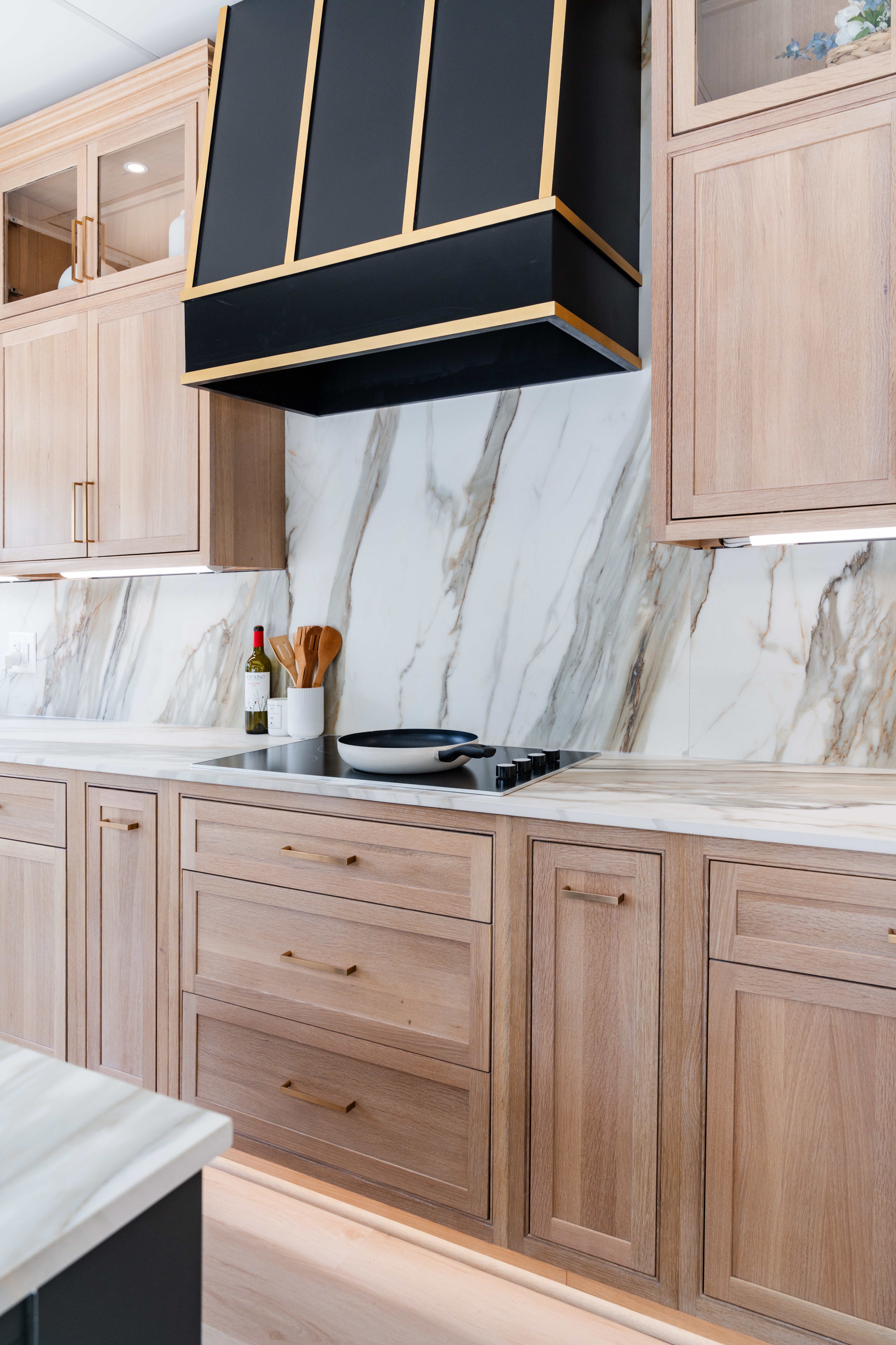 Modern kitchen with light wood cabinets, marble backsplash and countertop, a black and gold range hood, and a bottle of olive oil, utensils, and a pan on the stovetop—ideal inspiration for your next Maryland remodeling project.