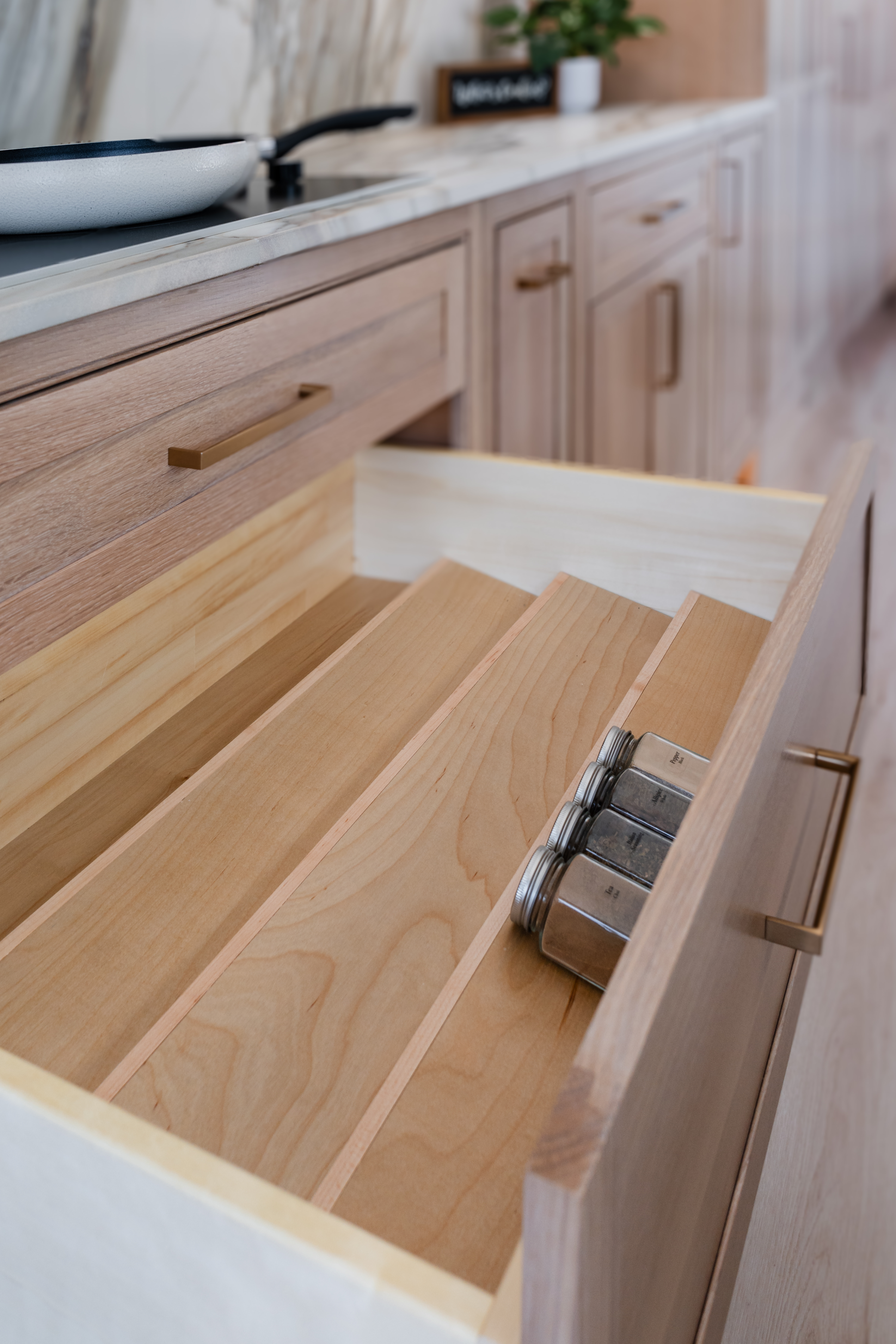 An open wooden kitchen drawer with angled dividers holds four glass spice jars in a modern Maryland kitchen, perfect inspiration for any remodeler seeking light wood cabinetry and a marble countertop.