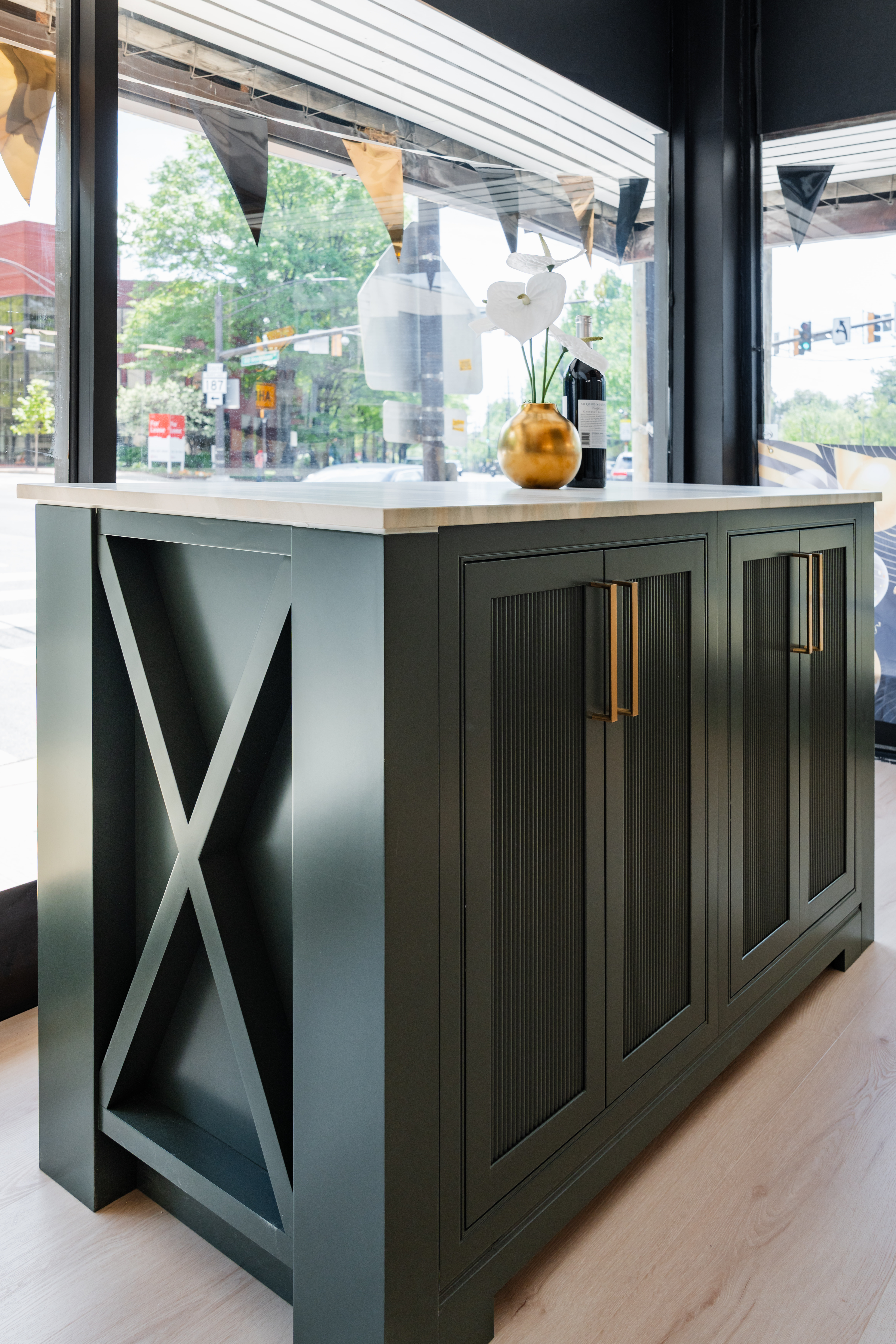 A modern green island cabinet with gold handles and a white countertop stands by large windows, perfect for a stylish DC remodeling. A small vase with white flowers decorates the top as sunlight streams in from outside.