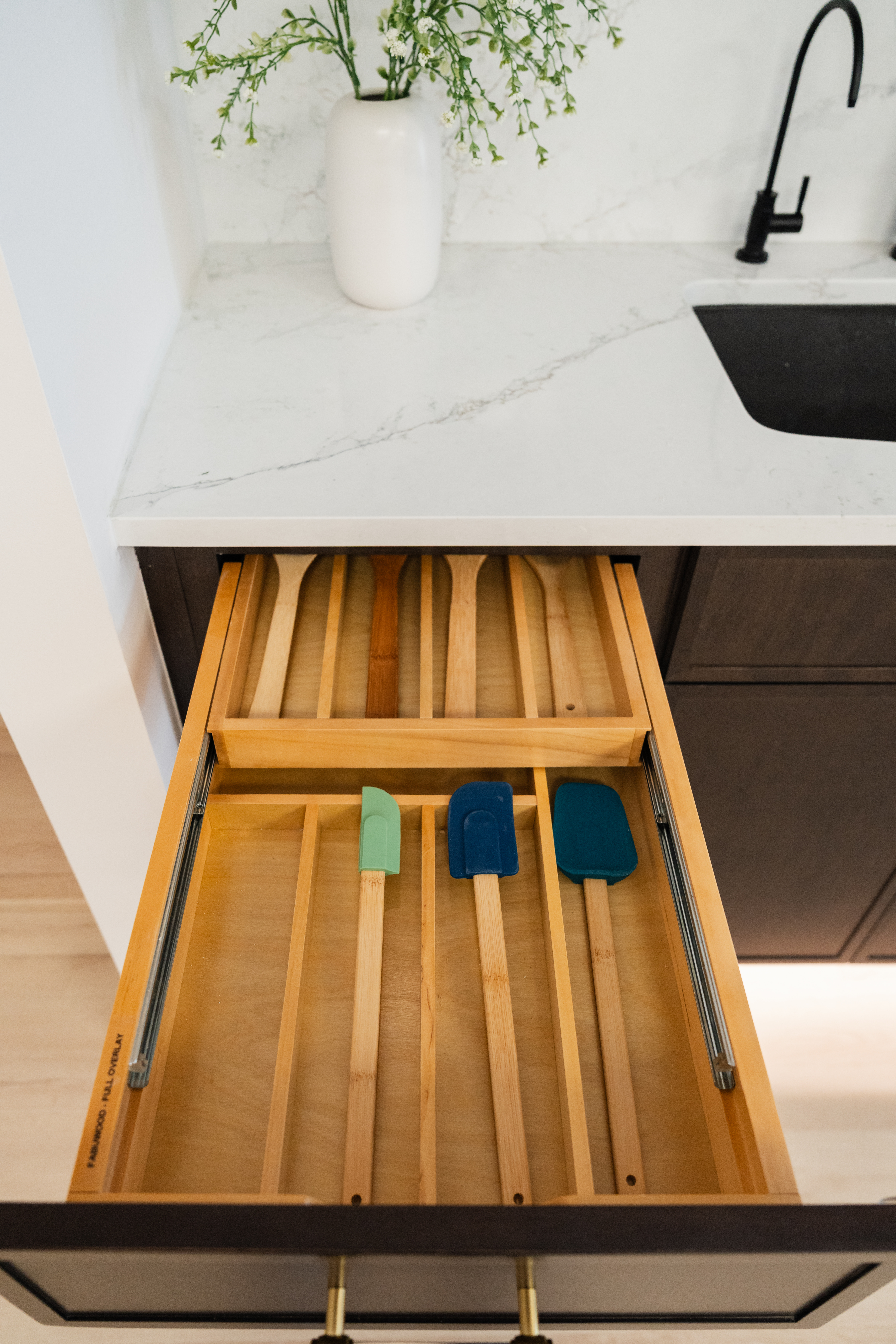 A kitchen drawer with wooden dividers holds wooden spoons and colorful silicone spatulas beneath a white countertop, expertly organized by a kitchen remodeler; above, a white vase of green flowers and a black faucet complete the scene.