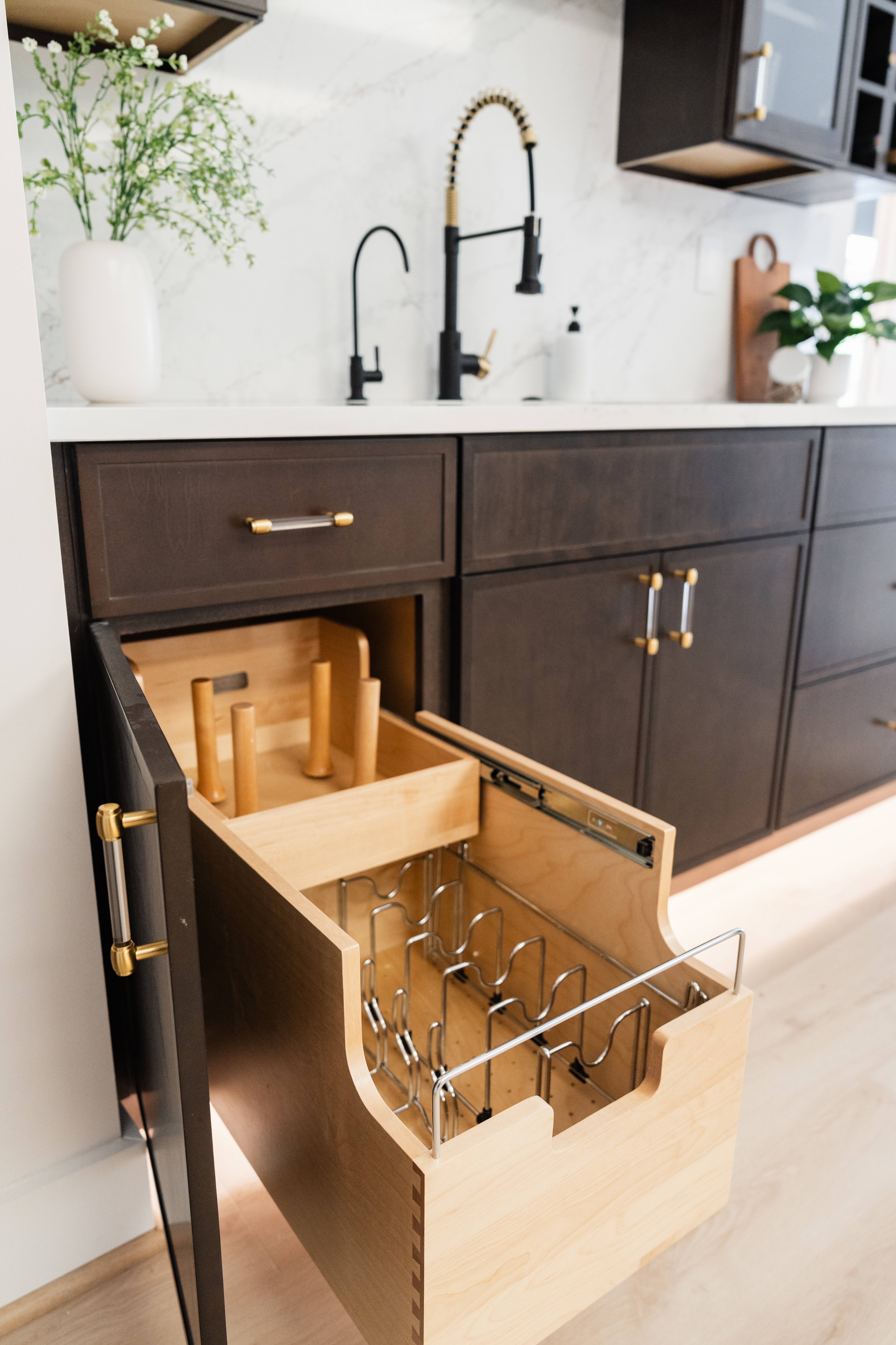 A modern kitchen by a top DC Kitchen Remodeler features dark cabinets with gold handles, plus an open pull-out drawer containing organized wooden knife slots and a metal rack for cutting boards or baking sheets.