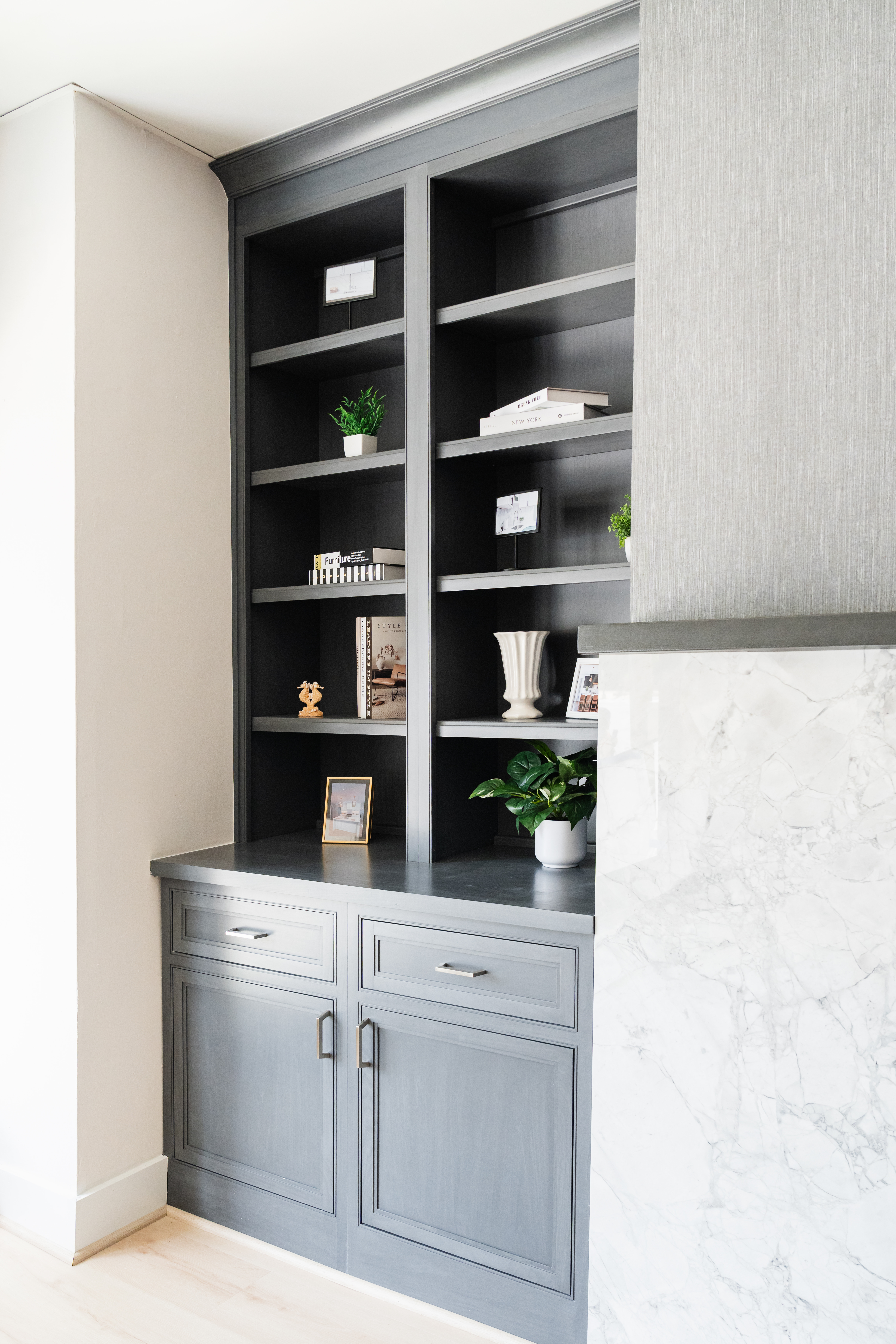 A built-in gray bookshelf with cabinets below, decorated with potted plants, framed photos, books, and vases, stands next to a mantel with white marble—perfect inspiration for any Maryland kitchen remodeler.