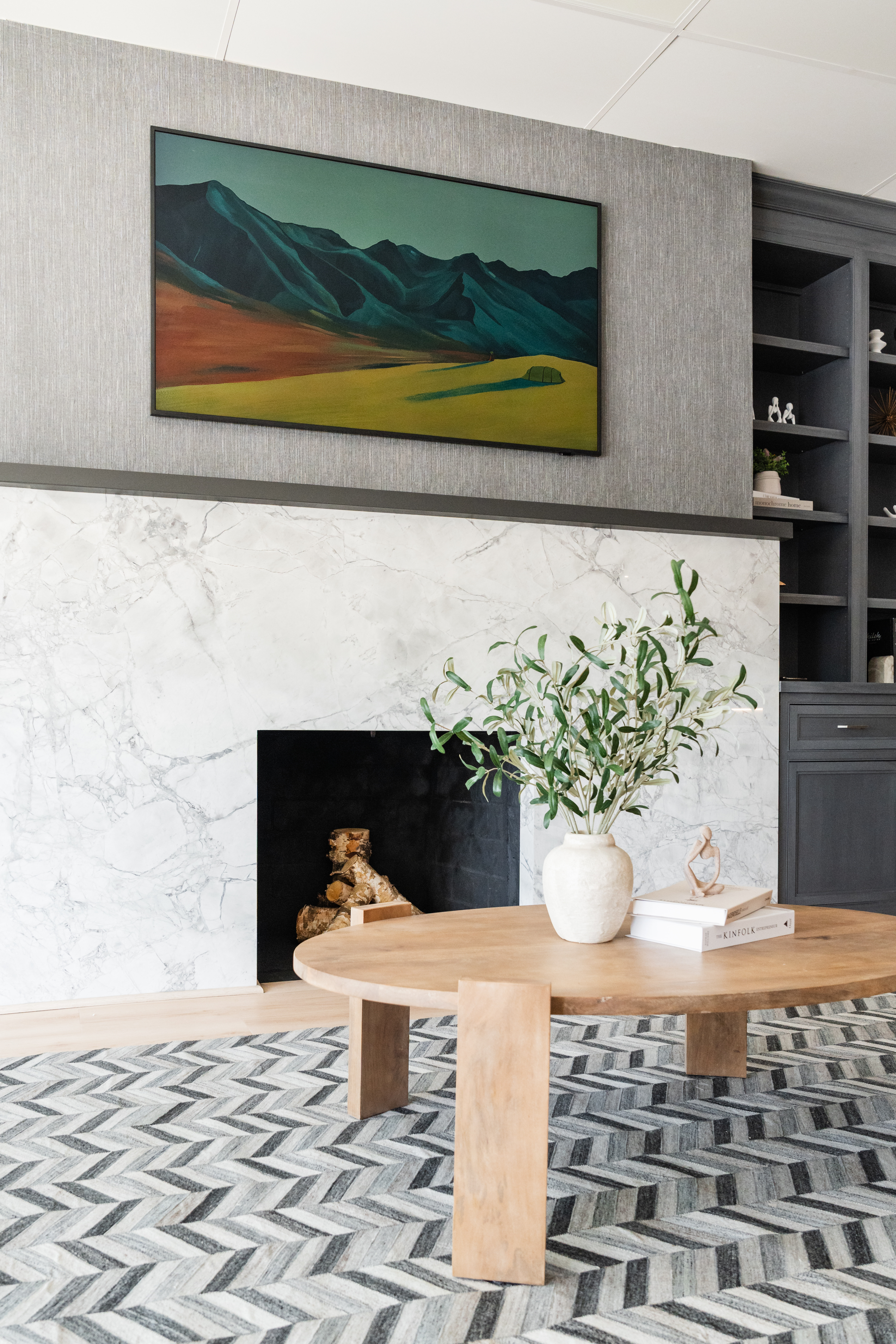 A modern living room, perfect for Maryland homes, features a round wooden coffee table, a white vase with green branches, a marble fireplace, built-in gray shelves, and a landscape painting of mountains and fields above the mantel.