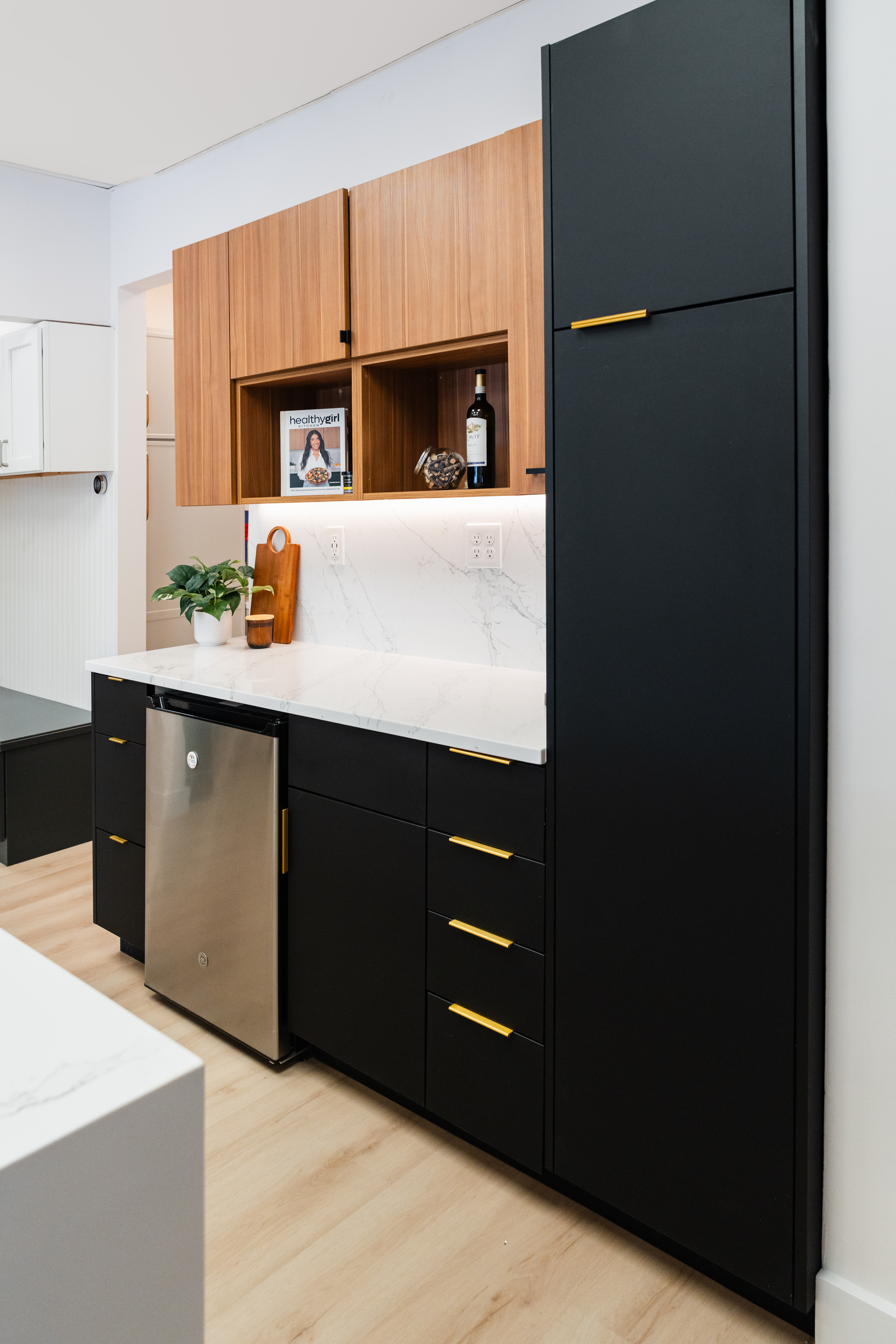 Modern kitchen by a skilled kitchen remodeler in DC, featuring black lower cabinets with gold handles, wood uppers, white marble surfaces, built-in dishwasher, potted plant, and minimal decor against light walls and floors.