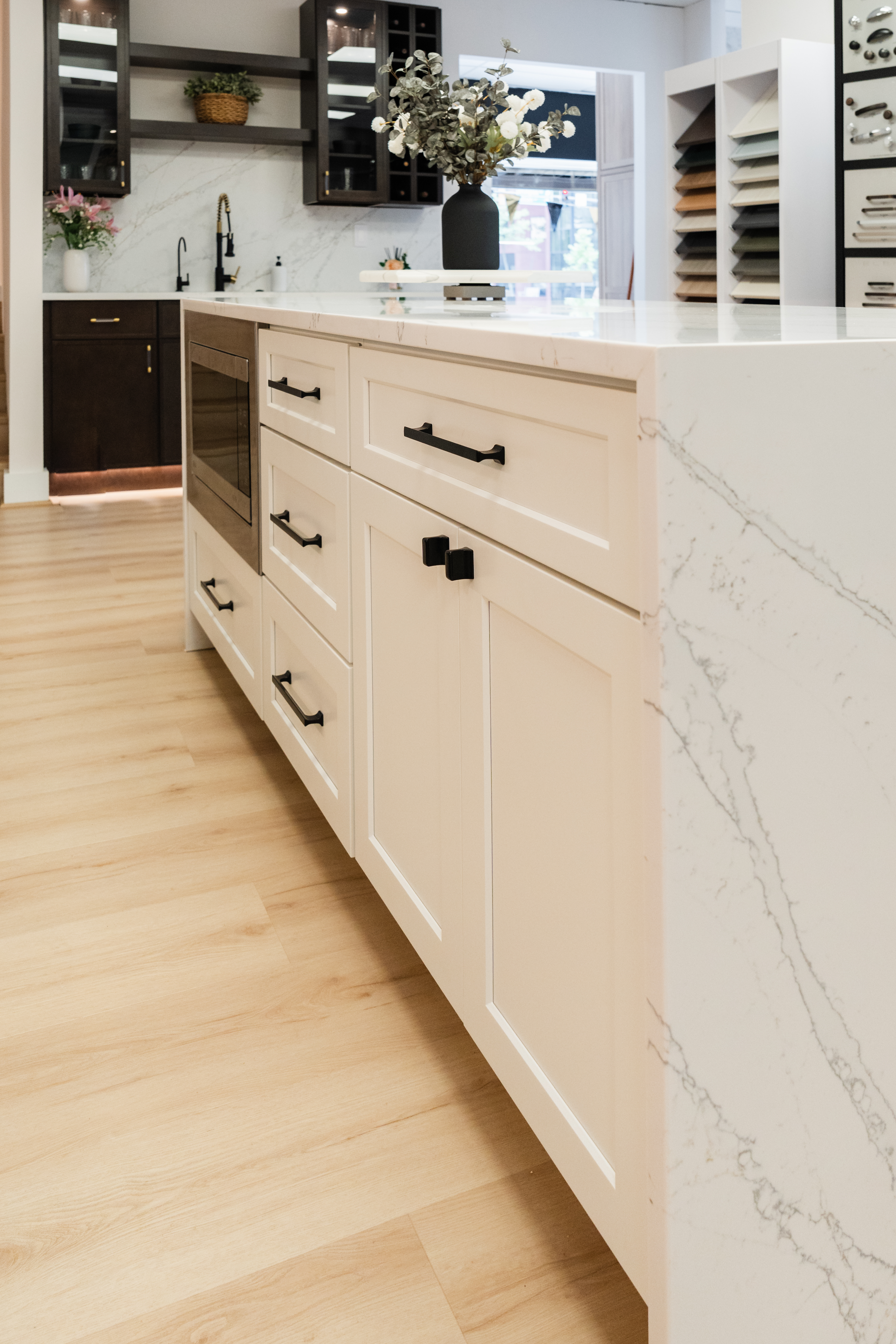 Modern kitchen island with white marble countertop, light wood floors, black hardware on drawers and cabinets, and a vase with white flowers. Open shelves and cabinets showcase the skill of a Maryland kitchen remodeler.