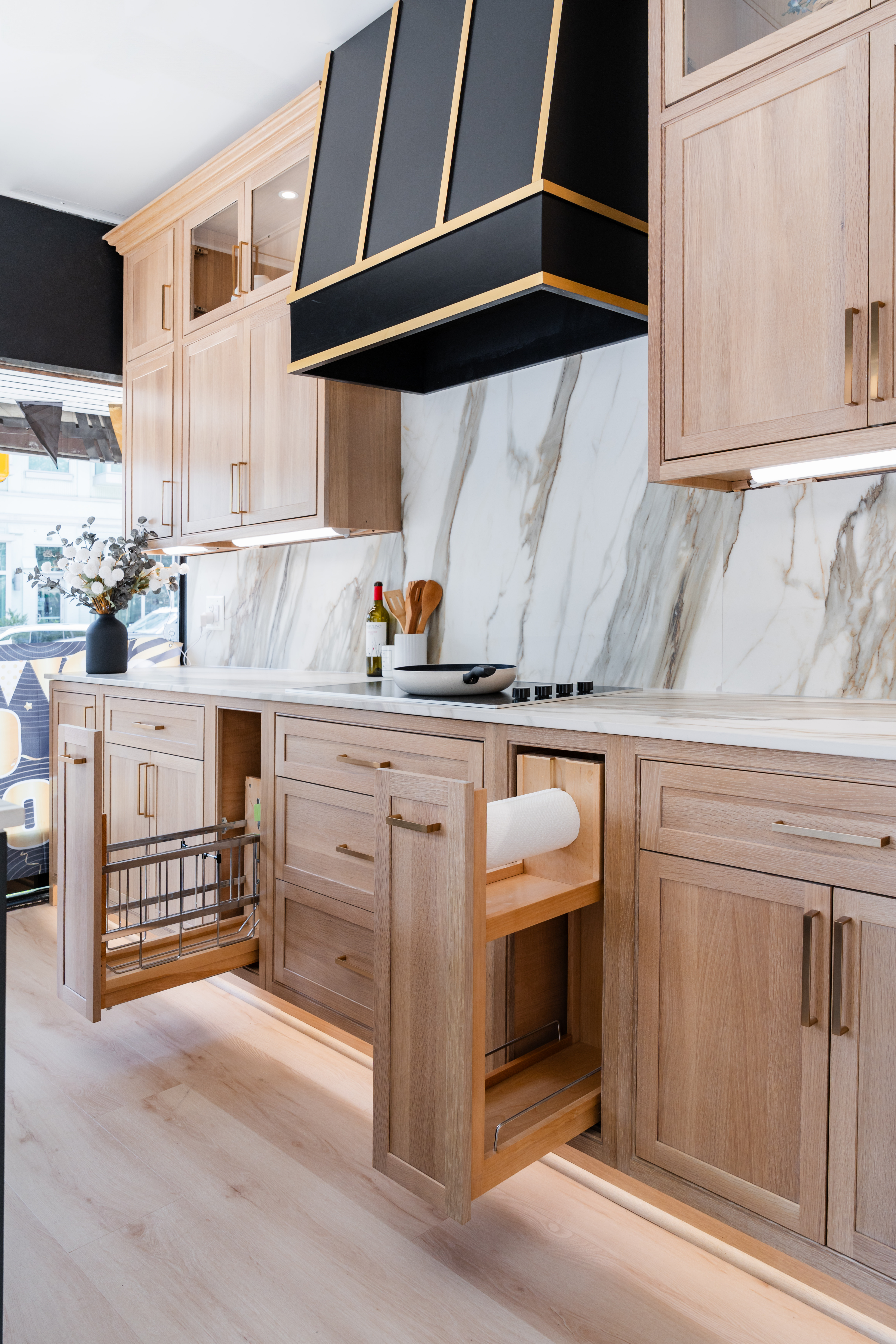 A modern kitchen, recently remodeled, features light wood cabinets, a marble backsplash, a black and gold range hood, built-in pull-out storage, and under-cabinet lighting. A vase with flowers and bottles sit on the counter.